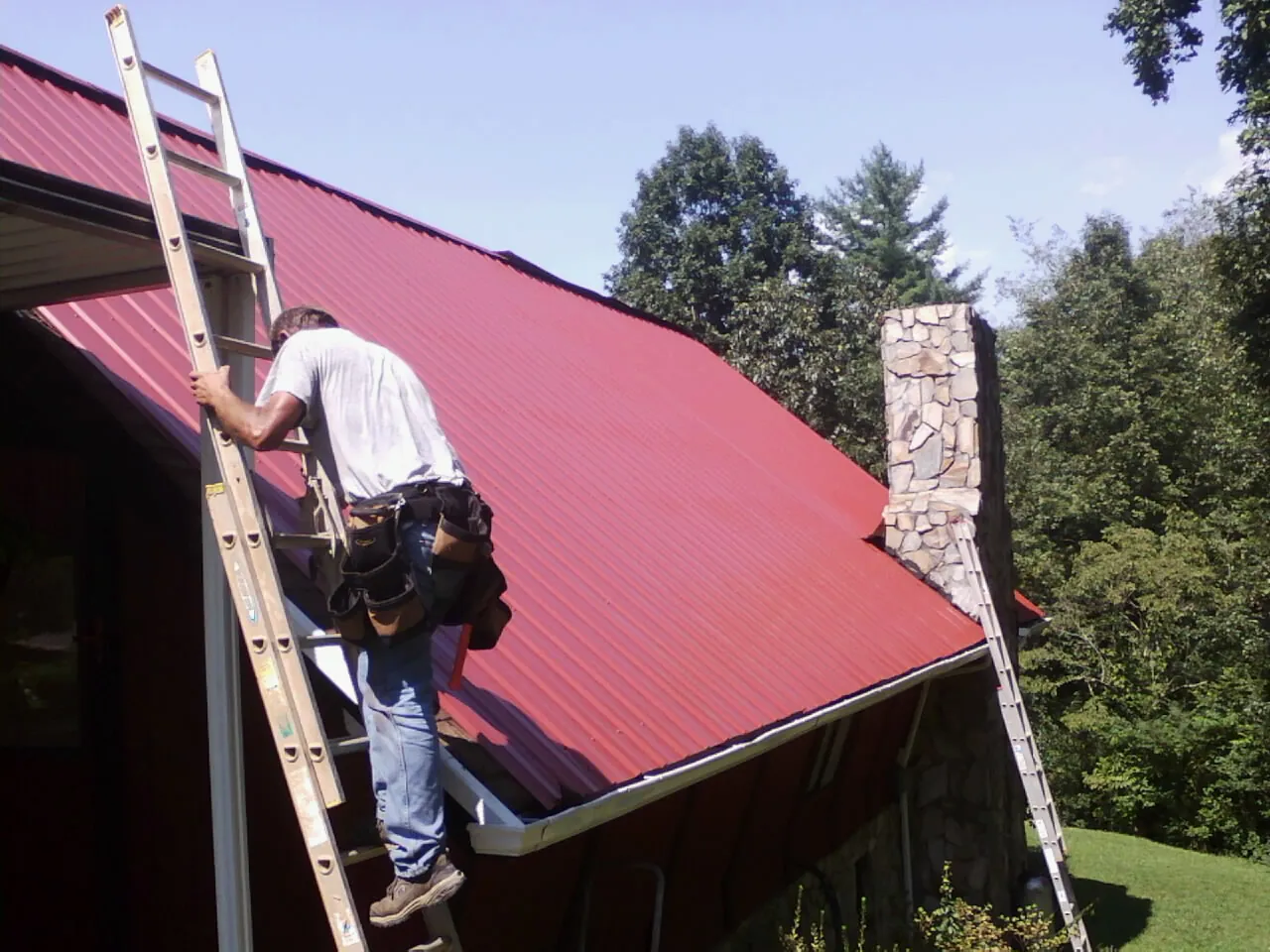 Roofer on a ladder beside a red metal roof and stone chimney — Soffit & Fascia Repair by Roofing Brothers in West Elizabeth